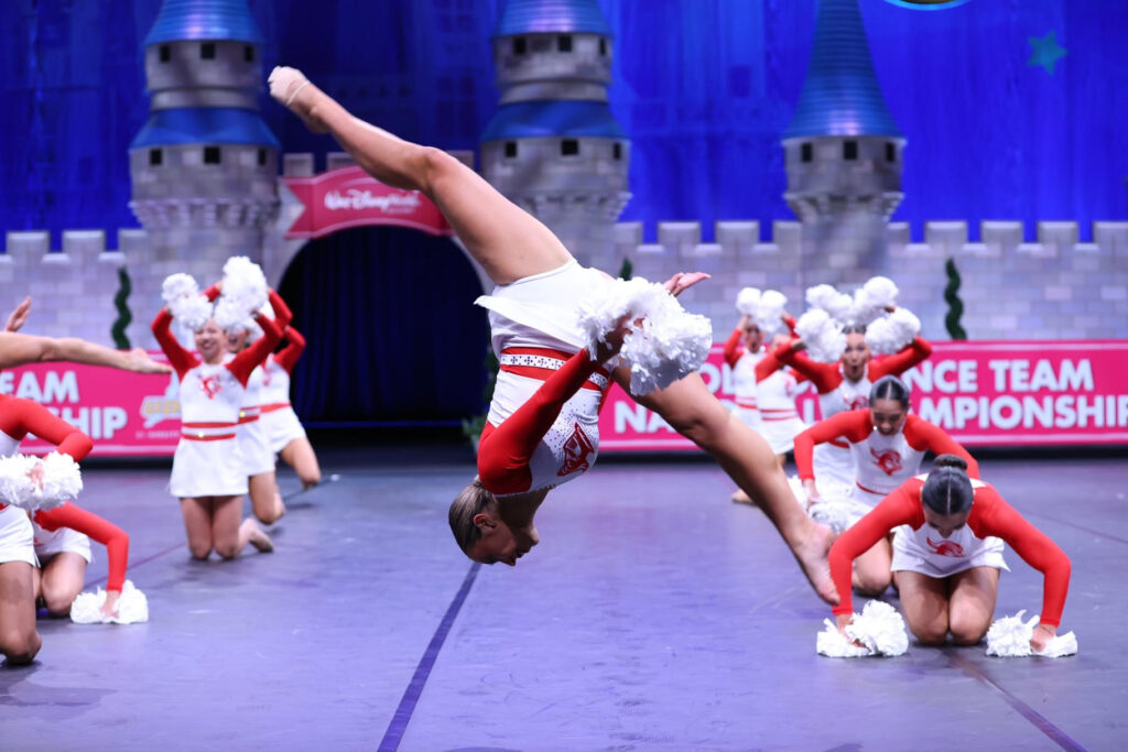 a dancer holding white poms and wearing a red and white uniform mid-air while performing a front aerial 
