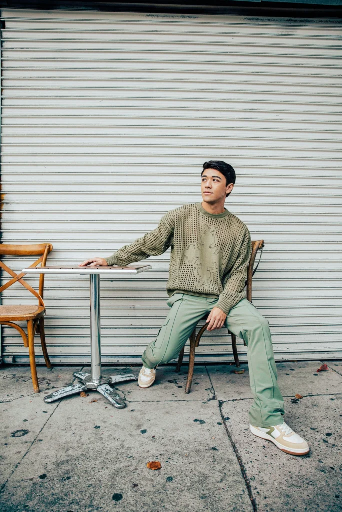 Kyle Hanagami, a young Asian man, sits at a café table set before a metal garage door. He looks thoughtfully to a corner while his right hand rests casually on a small table to his side. His feet are situated as though he could leap to his feet any instant. He wears a green mesh sweater and lighter green cargo pants.