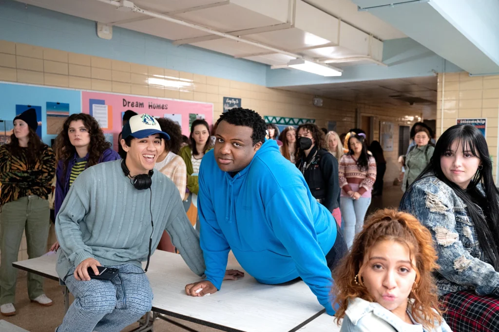 Kyle Hanagami perches on a cafeteria lunch table and cheeses at the camera. Beside him, Jaquel Spivey leans forward on both hands with a sardonic expression. Hanagami has a colorful ball cap on his head and headphones draped around his neck.