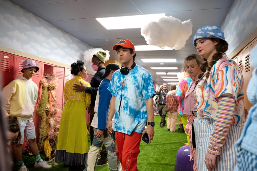 Kyle Hanagami walks through a film set designed to look like a high school hallway, but with grass between the lockers and faux clouds hanging from the ceiling. His expression is intent and focused. He has a set of headphones draped around his neck. Dozens of cast and crew members stand aside or are occupied with their own conversations.