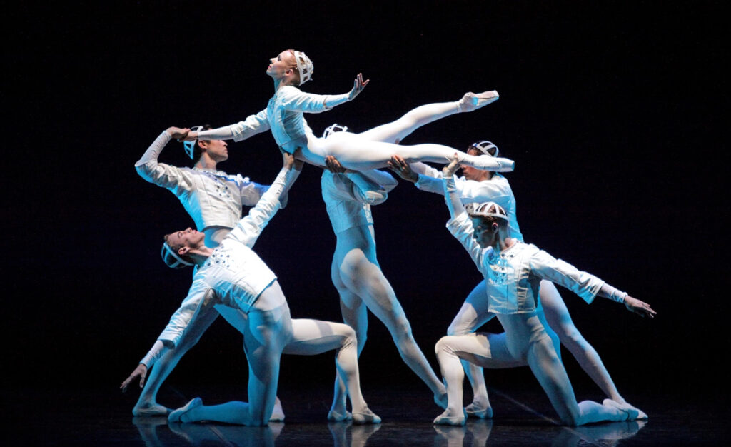 A female dancer is lifted horizontal to the floor, legs extended behind her and upper back arching up so she can look to the sky. She is supported by five male dancers, two kneeling downstage and three just upstage of her. All are dressed in white tights, matching long-sleeved tunics, and head coverings that have a futuristic feel.