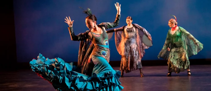 A flamenco dancer in a costume evocative of a peacock flicks her teal ruffled skirt behind her hands raised above the shoulder. In the background, two other dancers move through wide stances.