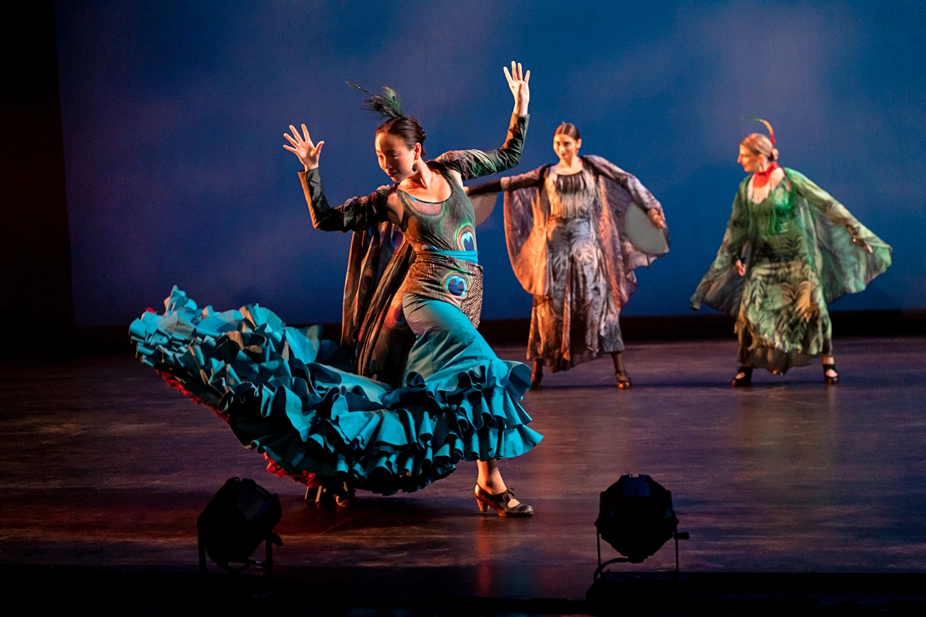 A flamenco dancer in a costume evocative of a peacock flicks her teal ruffled skirt behind her hands raised above the shoulder. In the background, two other dancers move through wide stances.