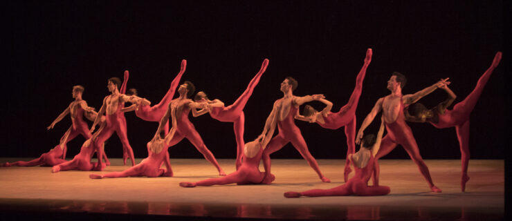There are four clusters of three dancers, all wearing red unitards. In each cluster, a male dancer lunges with one arm extended side for a female dancer to hold as she penchés, ducking her head below the supporting arm. Backs to the audience, the other women are seated before the men, heads resting against the men's bent knees.