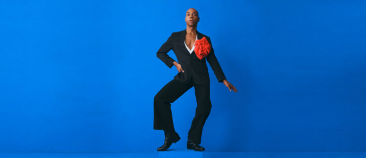 Shamel Pitts poses against a medium blue background. He wears a dark suit over a white shirt with a deep V and a large red handkerchief shaped into a flower at his lapel. He sits into one hip, legs turning out to an almost first position as he bevels one foot. That hand rests on his hip, while the other extends lightly out to the side at a downward angle.