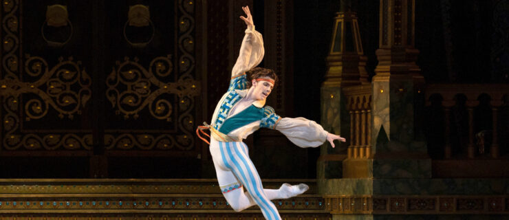 Jake Roxander is captured midair, arching back as he jumps in an overcrossed retiré back position. His costume features blue and white stripes down one leg and teal patterns on his puffy-sleeved tunic. The set behind him evokes an opulent ballroom.
