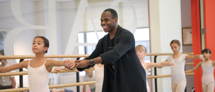 McDaniel, a dark-skinned man wearing black practice clothes, smiles as he corrects the arm placement of a young student in a ballet classroom.