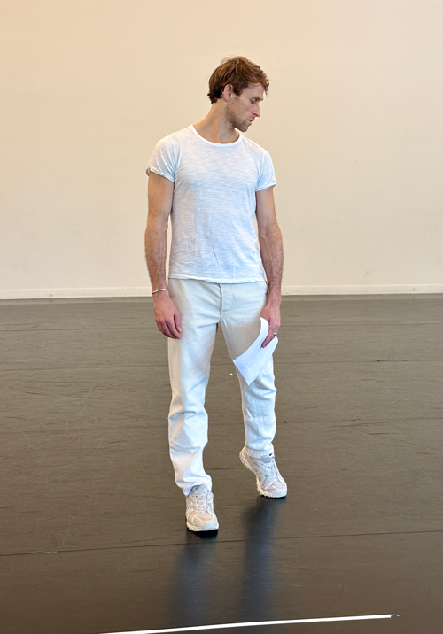 Danchig-Waring, wearing white practice clothes and sneakers, stands in a dance studio, holding a collection of papers in his left hand and looking down over his left shoulder.
