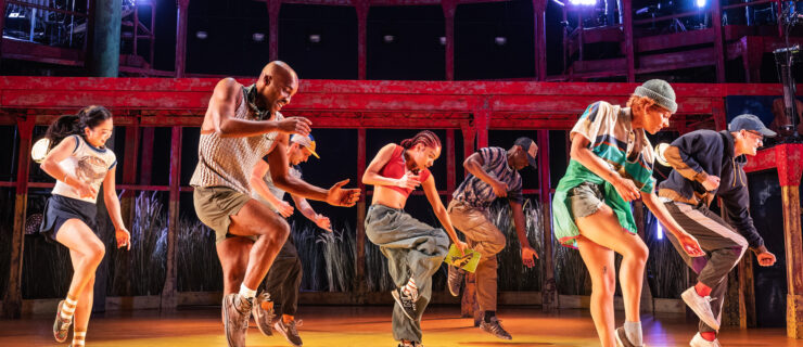 A collection of dancers in colorful street clothes and sneakers are captured onstage mid-jump, with their right knees slightly bent and their arms floating loosely. In the background, red scaffolding looms above the golden floor.