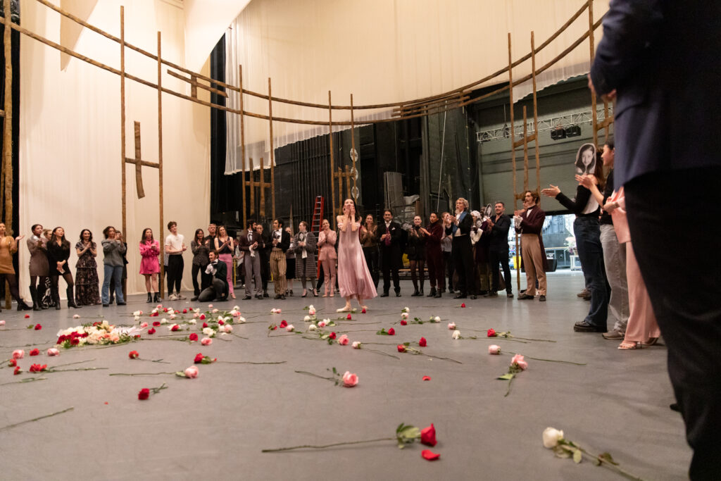 a female dancer in center with roses all over the ground being applauded by a group of onlookers 