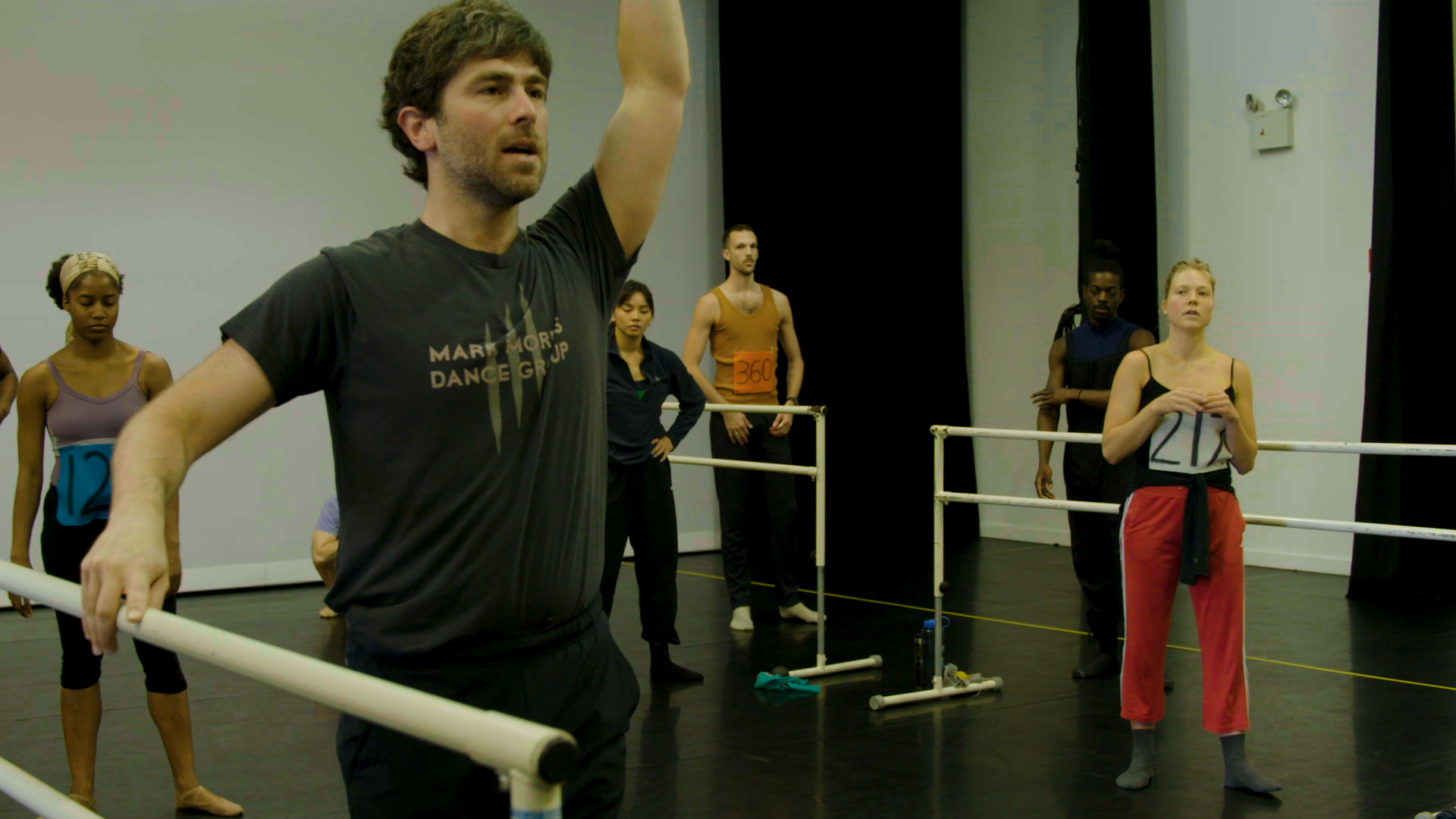 Several dancers with numbers pinned to their rehearsal clothing stand in a dance studio, watching Black—wearing a Mark Morris Dance Group t-shirt—demonstrate a combination. Black's right hand rests on a barre and his left is raised above his head.
