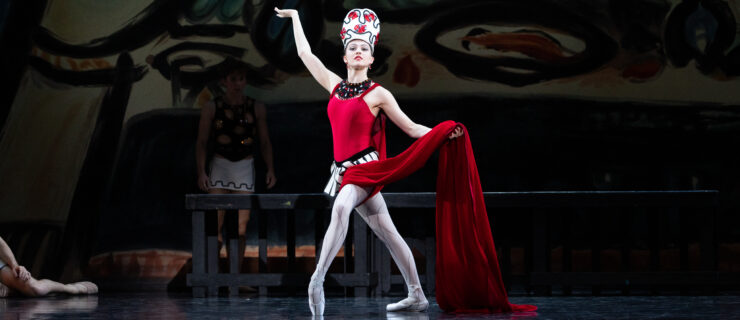 Sydney Dolan onstage as the Siren in Balanchine's Prodigal Son. She poses center stage in a fourth position lunge, front foot in forced arch on pointe. Her left hand holds the long red train of her costume at waist height; her right hand is raised to the side, palm up.